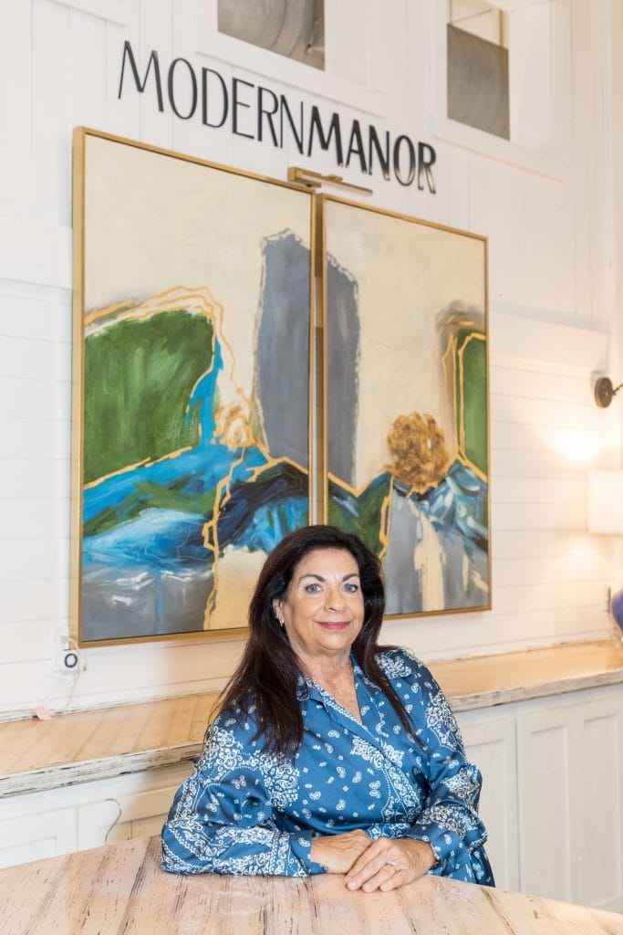 Modern Manor A woman in a blue patterned outfit sits at a wooden table in front of a large abstract painting and a sign that reads "MODERN MANOR.
